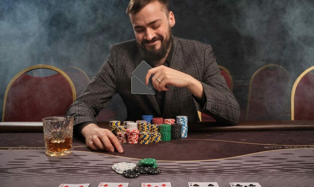 A man seated at a poker table, wearing a smile as he examines his cards. Poker chips are neatly stacked in front of him on the table.