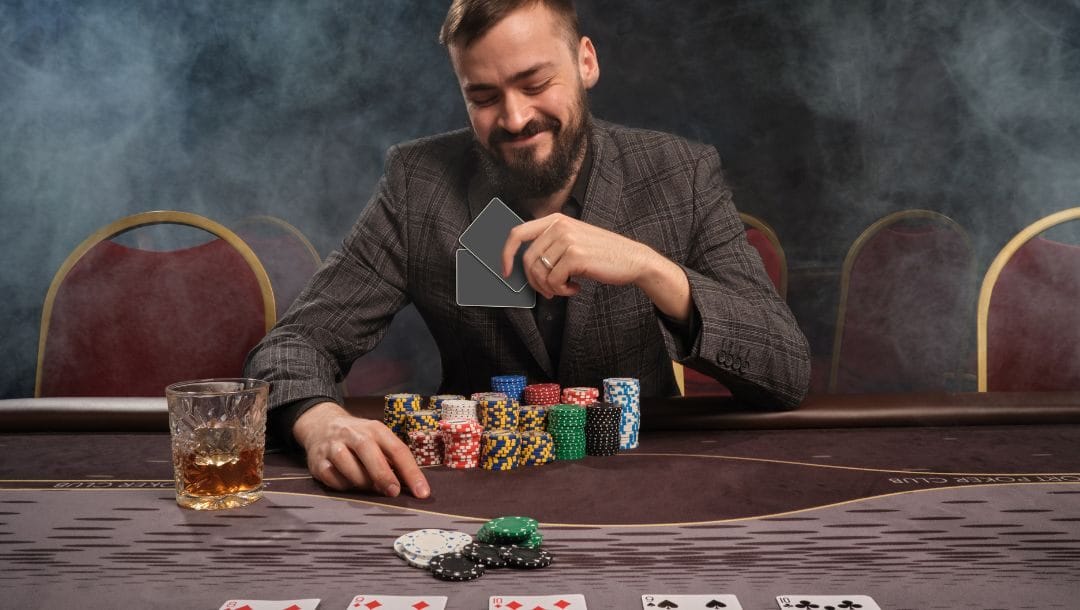 A man seated at a poker table, wearing a smile as he examines his cards. Poker chips are neatly stacked in front of him on the table.