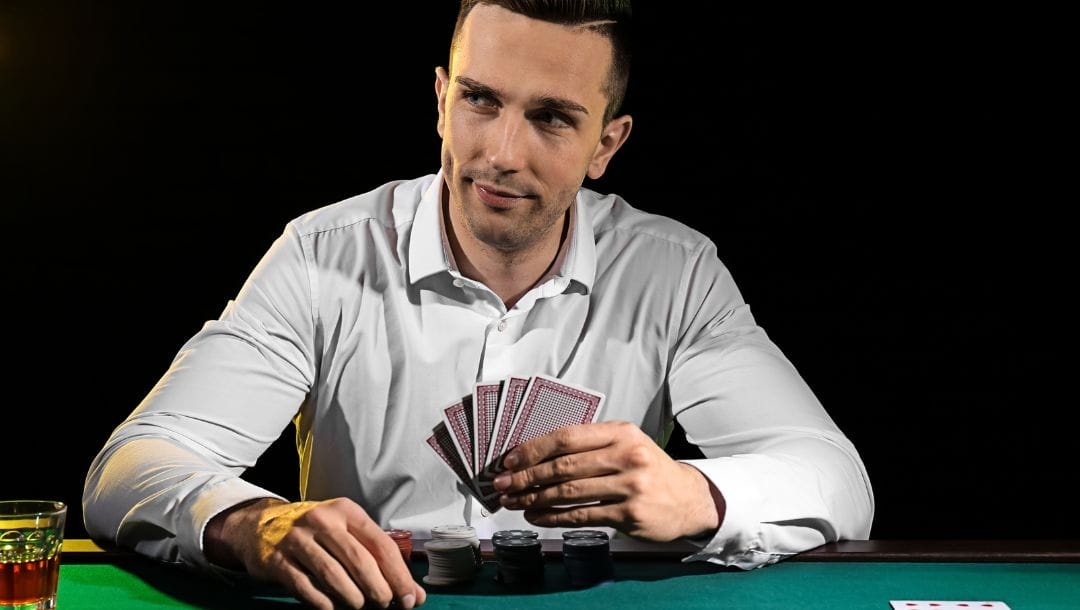 A man sitting at a poker table, holding poker cards, smirking and glancing to the left. Poker chips are neatly stacked in front of him on the table.