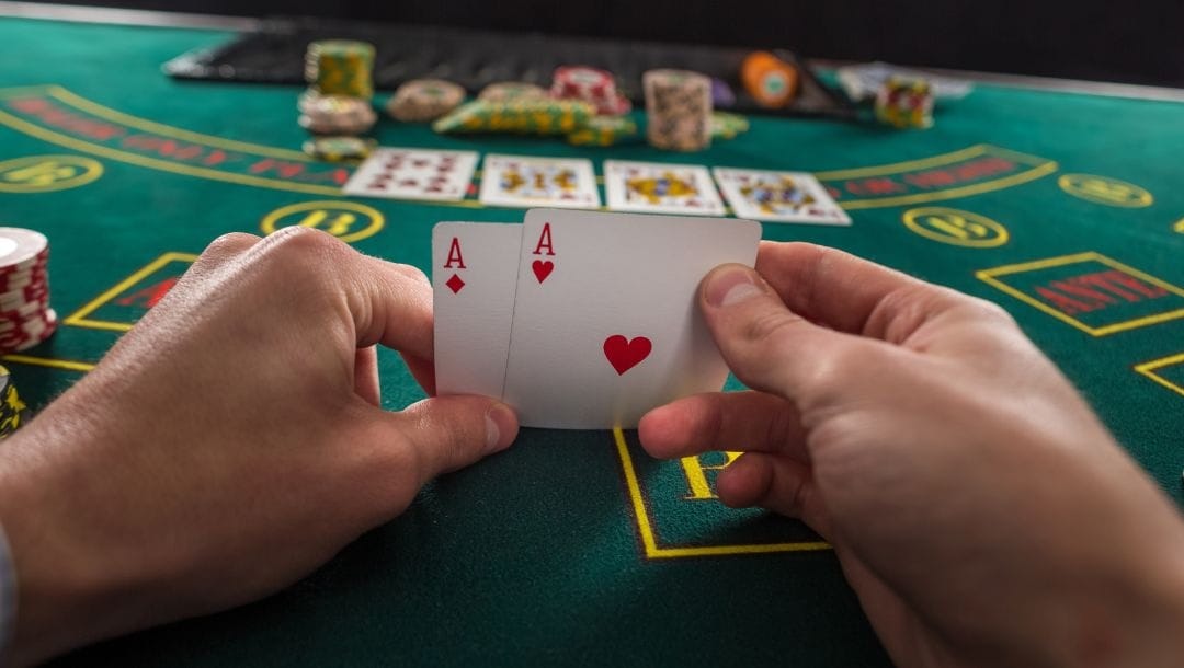 A close up of a poker player’s hands holding a pair of Aces in the diamond and hearts suits.