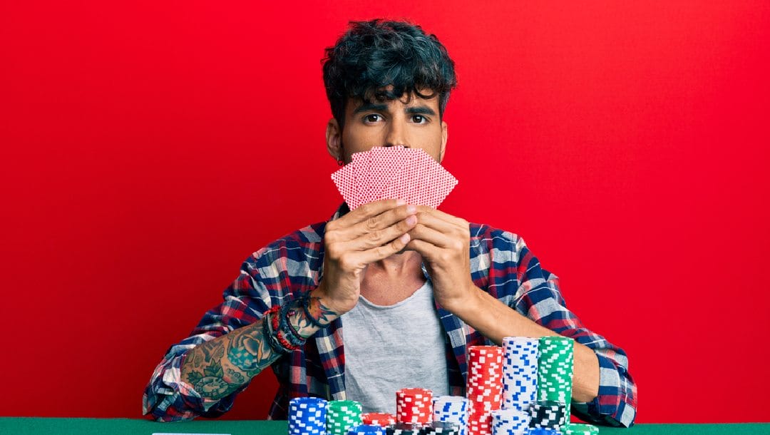A man holding cards in front of his mouth with poker chips in the foreground and a red wall in the background