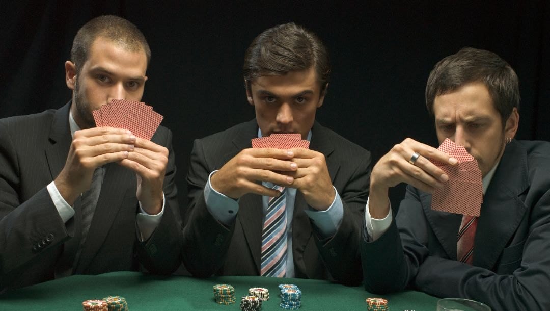 three men holding playing cards up covering their lower face at a poker table with poker chips on it