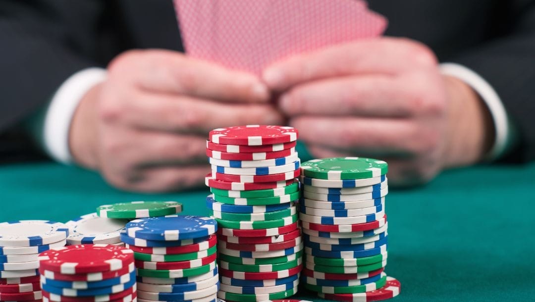stacks of multicolored poker chips on a green felt poker table with a man holding playing cards in the blurred background