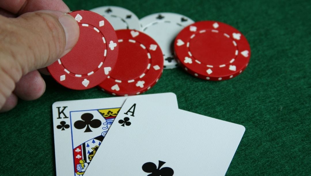 a close up of a person holding a red poker chip above two playing cards, an ace and king of clubs, on a green felt poker table with other red and white poker chips on it