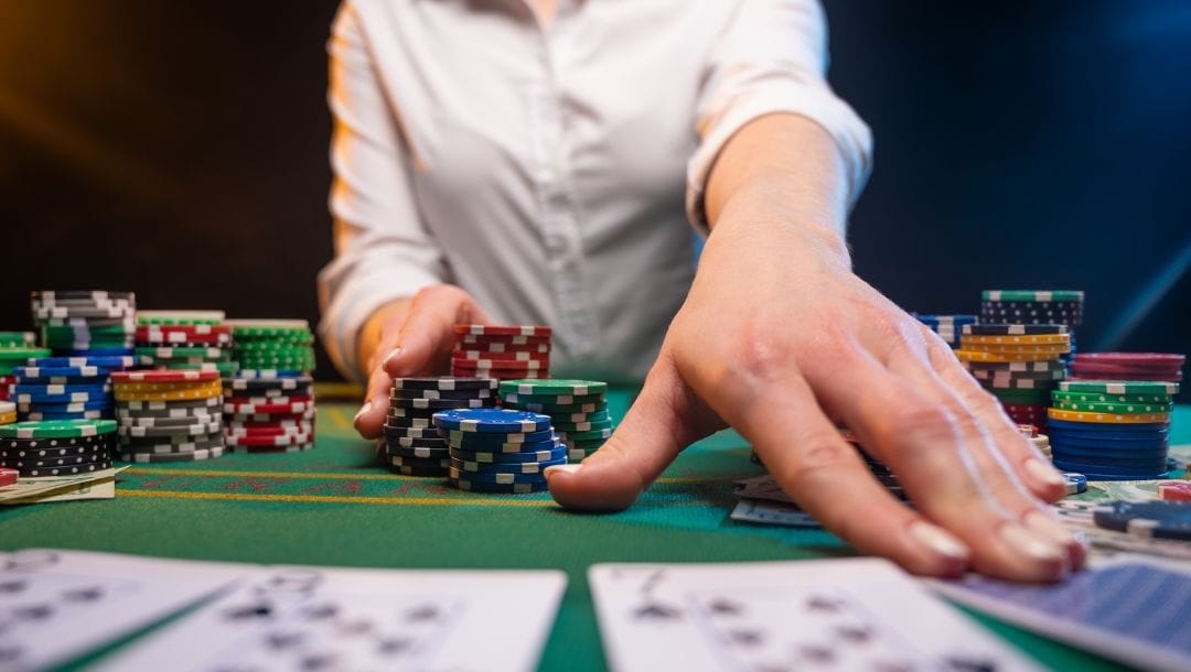 A neck-down photograph of a dealer sitting at a poker table, reaching forward to touch the playing cards closest to the camera. The dealer’s other hand rests on a large stack of poker chips.