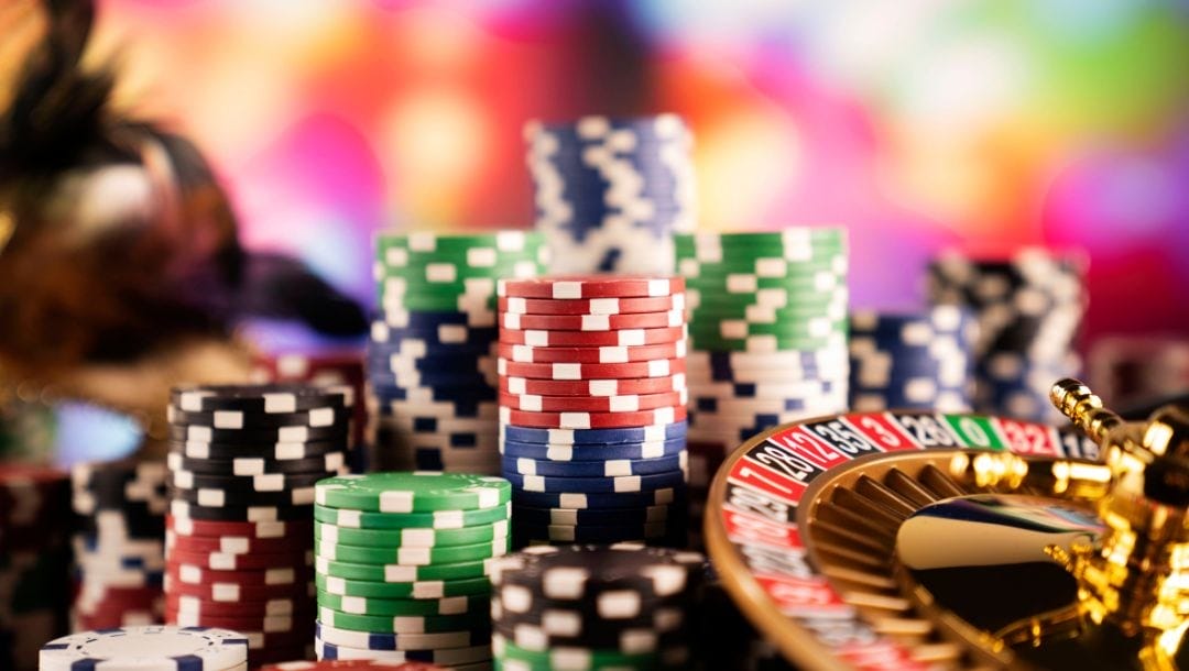 A full color image of poker chips of different values and colors, stacked next to a small, gold roulette wheel.
