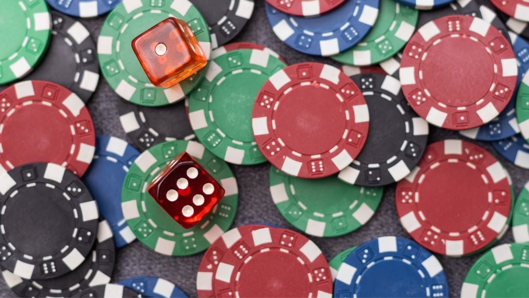 Two red six-sided dice sitting on top of green, red, blue and black poker chips that have been scattered across a black background.