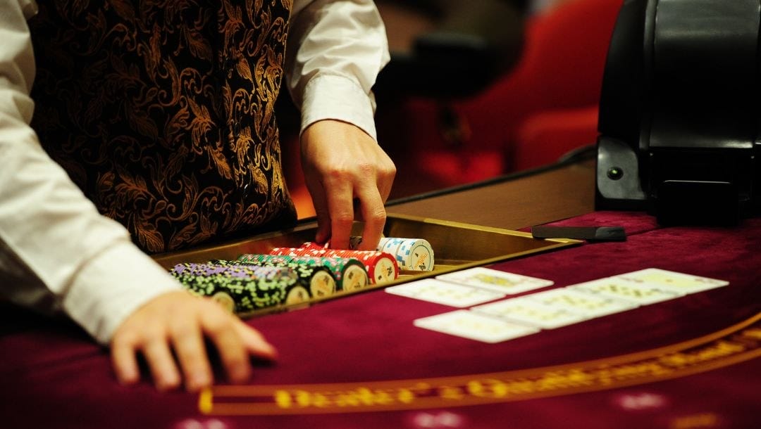 A closup of a poker dealer standing at the poker table with one hand resting on the poker chips and the other resting on the table.