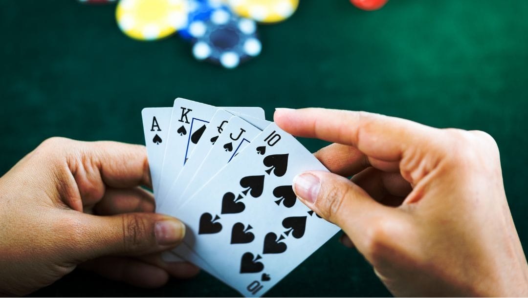 a close up of a person holding a royal flush of spade playing cards above a green felt poker table with some poker chips on it blurred in the background