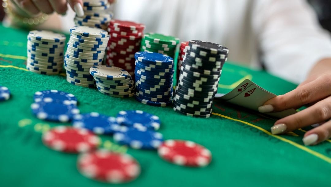 a person lifts their hole cards to reveal a pocket pair of ace playing cards on a green felt poker table next to stacks of multi colored poker chips with some poker chips blurred in the foreground
