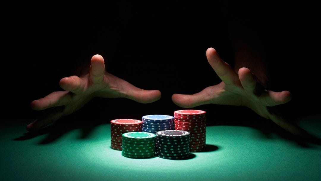 a pair of hands reaching out of the shadows to grab stacks of poker chips on a green felt poker table