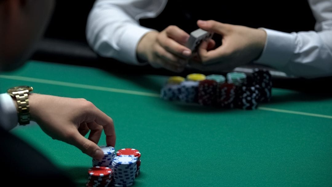 an over-the-shoulder view of a poker player sitting opposite a dealer at a poker table in a casino, both have stacks of poker chips in front of them and the dealer is holding a deck of playing cards