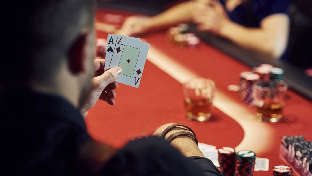 an over-the-shoulder view of a man looking at the two playing cards in his hand, a pocket pair of aces, while sitting at a poker table playing poker in a casino