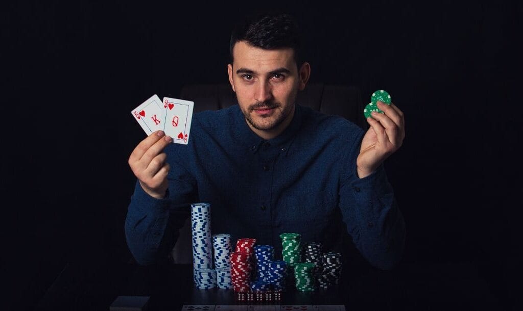 a man holding two playing cards and two green poker chips is sitting at a table that has stacks of poker chips on it