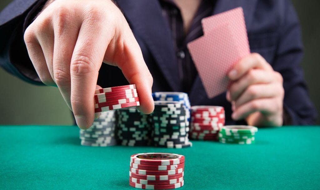 a man adding to a stack of red poker chips on a green felt poker table while holding two playing cards with poker chips stacked in front of him on the table