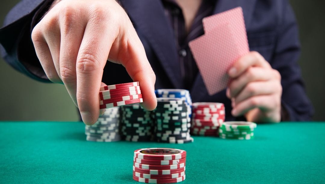 a man adding to a stack of red poker chips on a green felt poker table while holding two playing cards with poker chips stacked in front of him on the table