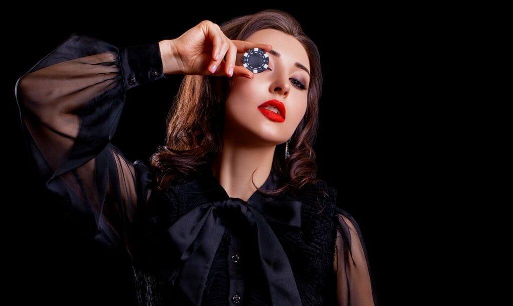 A woman in a black dress holds a casino chip up to her eye.