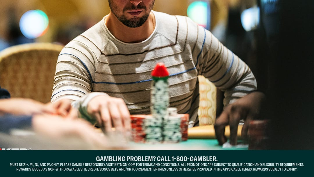 A focused poker player in a striped sweater sitting at a casino table, stacking a large pile of poker chips with a single red chip on top. Other players' hands are visible in the foreground.