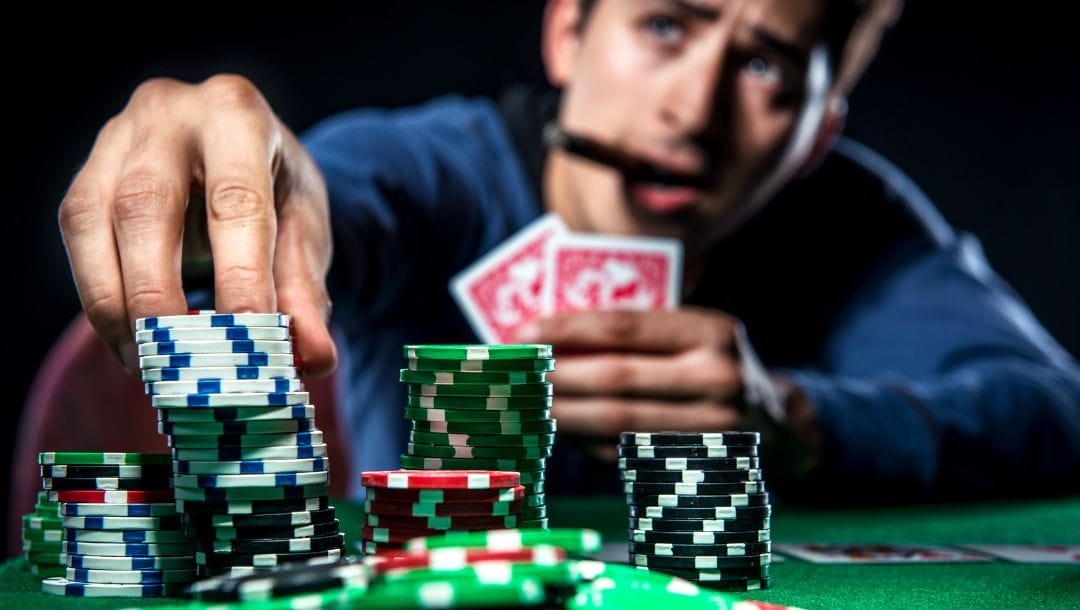 A man sitting at a poker table, holding two cards in his one hand, and placing casino chips with his other hand.