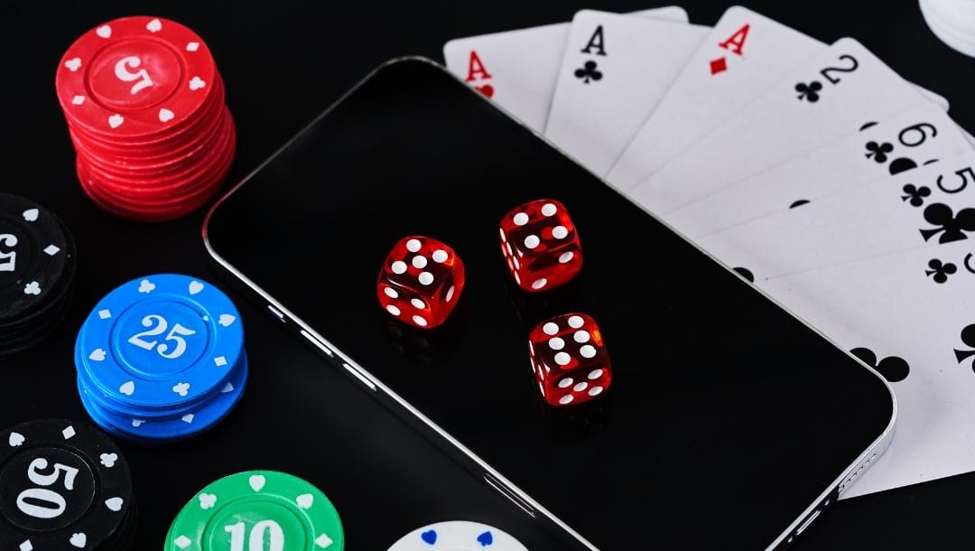 Three red six-sided dice on top of a cellphone with small stacks of poker chips and playing cards surrounding it.
