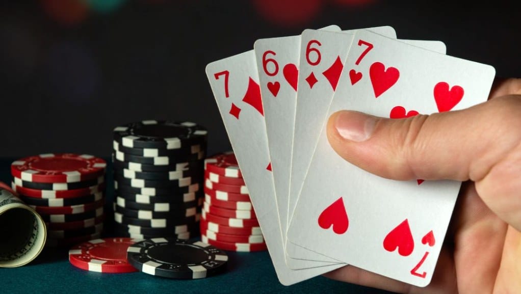 A closeup of a hand holding a pair of sixes and a pair of sevens with red and black poker chips and rolled up cash in the background.