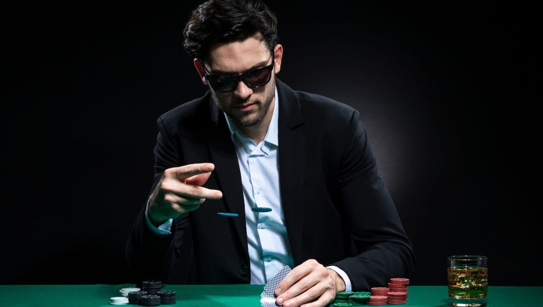 A man wearing a suit and sunglasses, seated at a poker table, with a stack of casino chips neatly arranged in front of him.