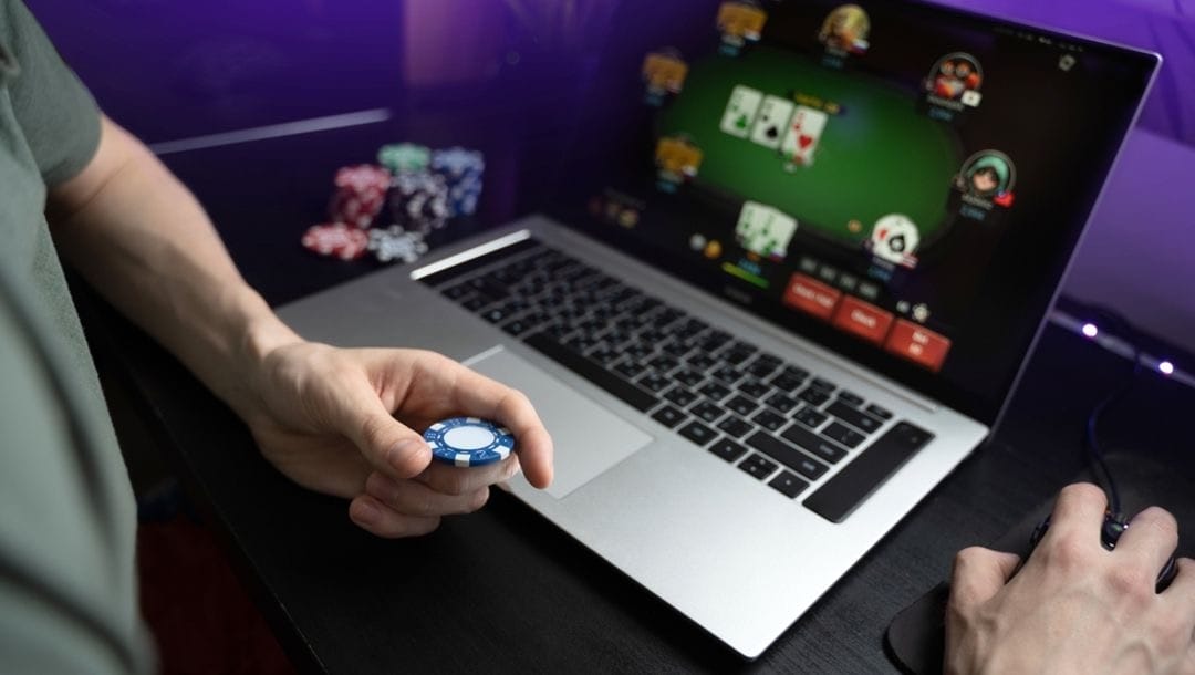 A man holding a casino chip playing poker on a laptop.