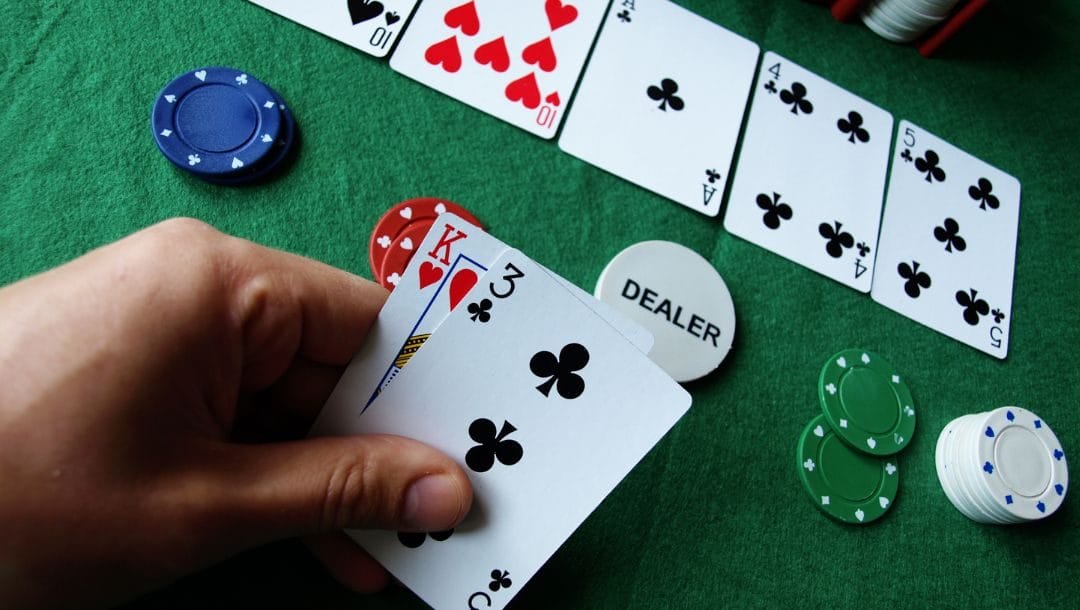 Hand displaying cards at a poker table, revealing a King of Hearts and a Three of Clubs, surrounded by additional cards and poker chips.