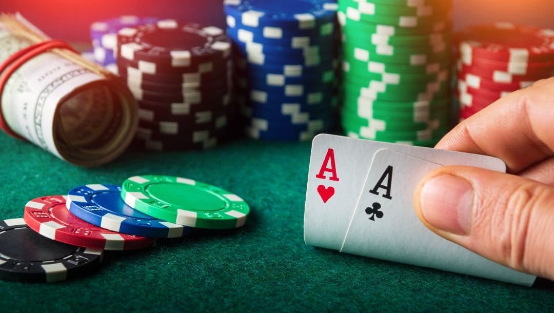 Hands revealing an Ace of Hearts, and an Ace of Clubs on a poker table, with a roll of money and poker chips neatly arranged nearby.