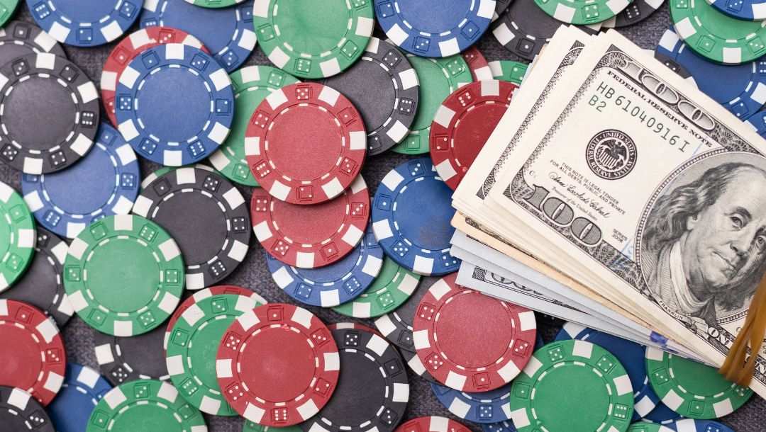 A birds-eye view of different color poker chips scattered across a black background with a stack of $100 bills being held together by a rubber band sitting on top of them.