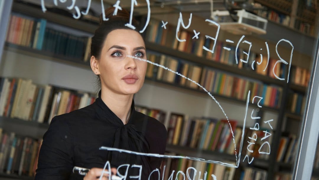 A person looking at a mathematical formula on a transparent board. There are shelves of books behind them.