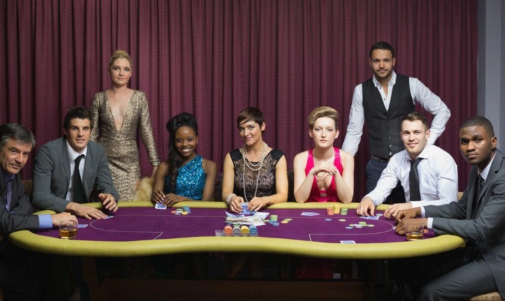 A well-dressed group of people sitting and standing around a poker table.