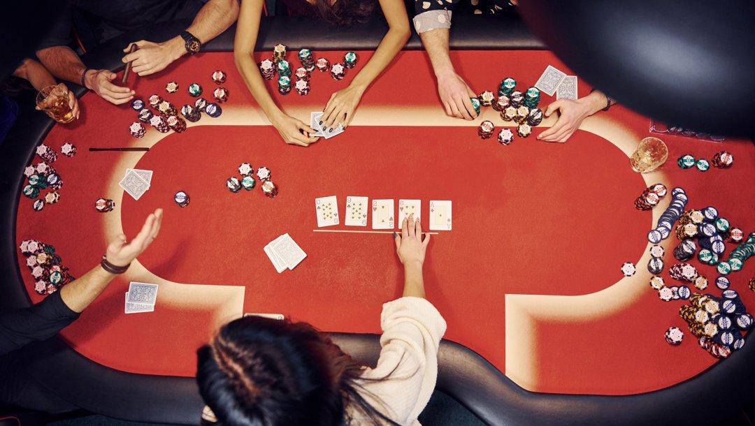 An aerial view of a red poker table. All five community cards are out and the dealer is engaging with one of the five players at the table.