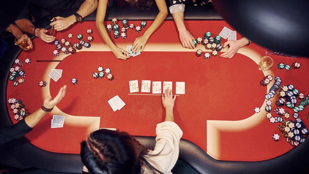 An aerial view of a red poker table. All five community cards are out, and the dealer is engaging with one of the five players at the table.