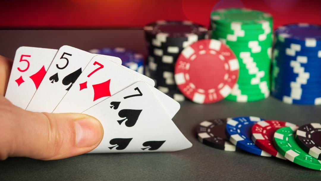 A PLO player checks their hand. They have a pair of 5s and a pair of 7s. There are small stacks of poker chips in the background.