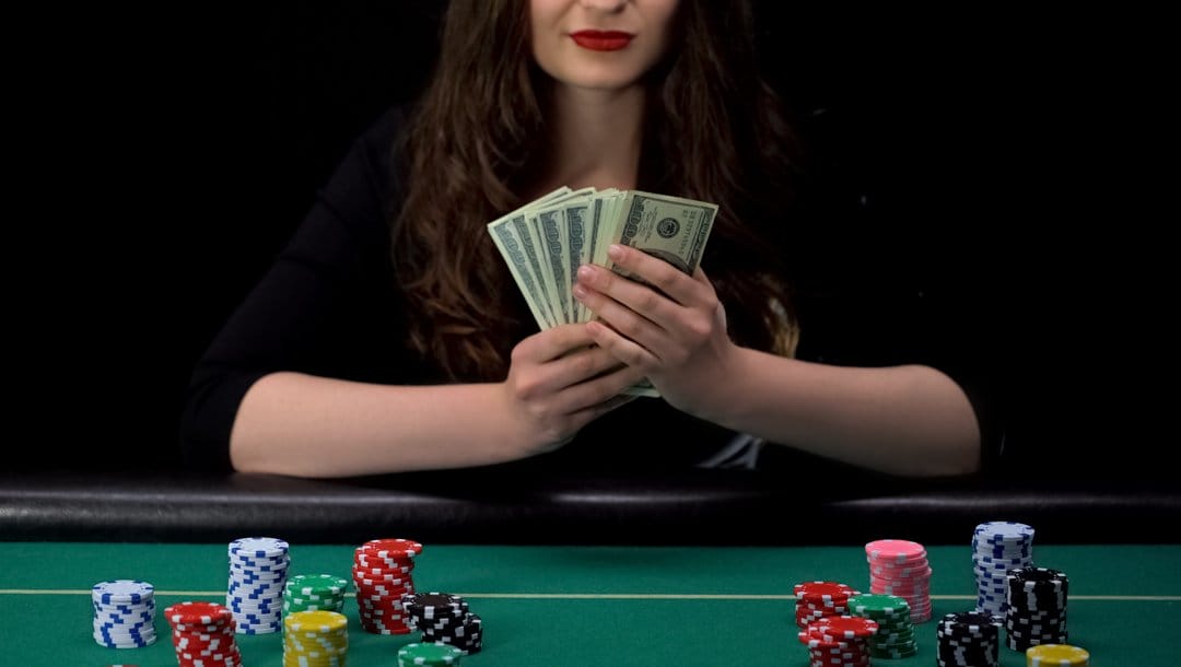 A player at a poker table holding $100 bills in their hand. There are stacks of poker chips on the table.