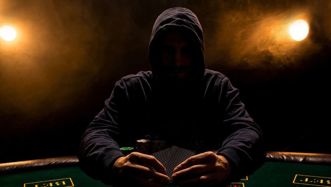 a mysterious figure of a man sitting at a poker table wearing a hoodie while holding playing cards