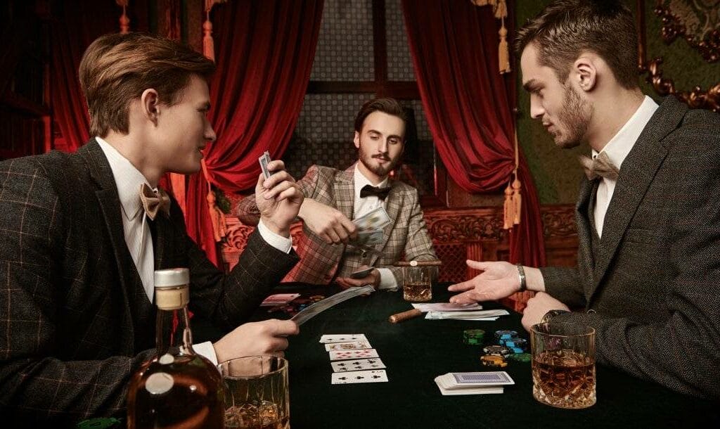 Three people in tweed suits playing poker in a Victorian-style room. There are drinks, cigars, playing cards, and poker chips on the table in front of them.