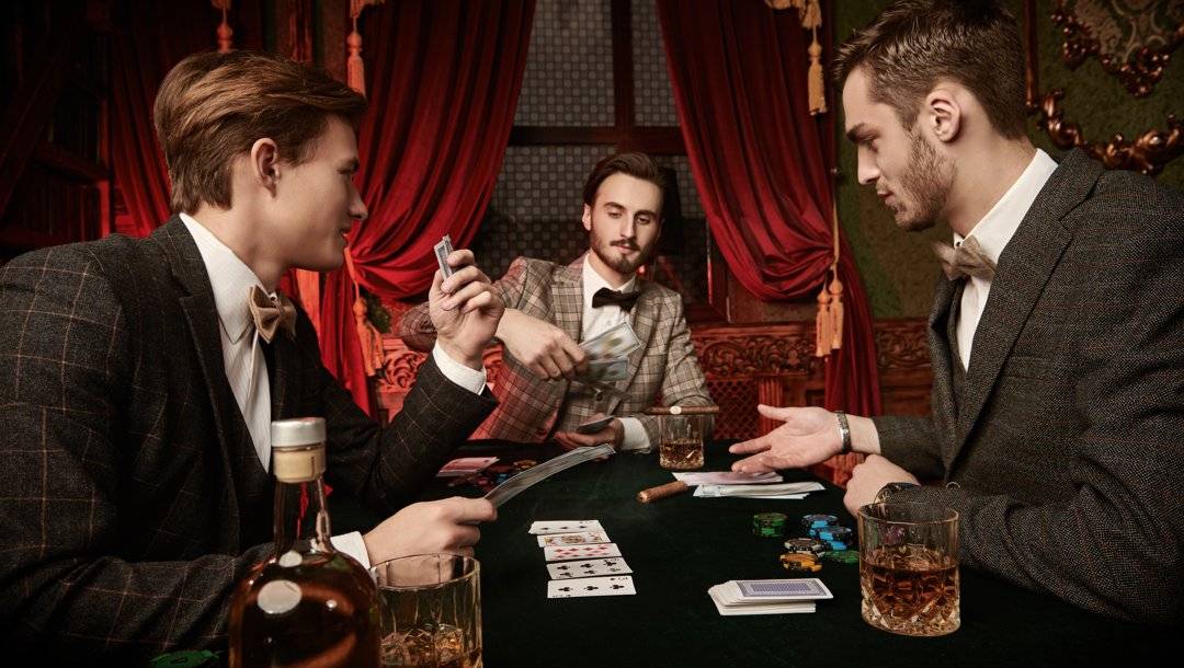 Three people in tweed suits playing poker in a Victorian-style room. There are drinks, cigars, playing cards, and poker chips on the table in front of them.
