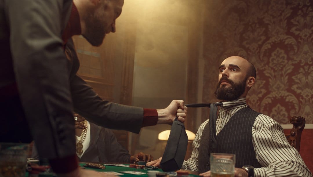 An angry poker player grabs another poker player’s tie and pulls them forward. Another player watches.
