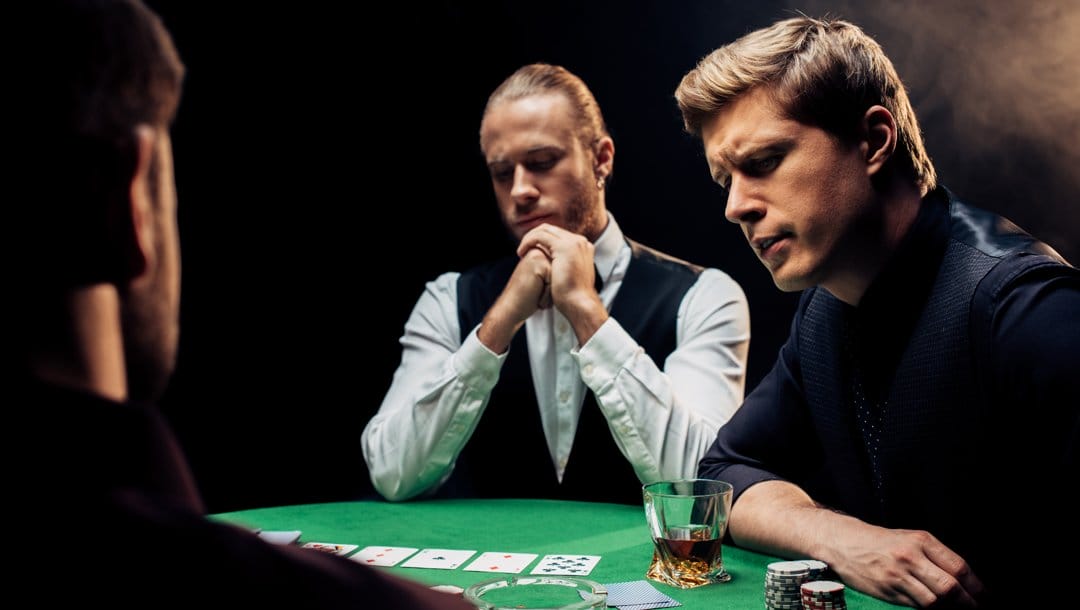 A poker player faces another player who looks concernedly at the poker table. The dealer is also looking at the cards on the table.