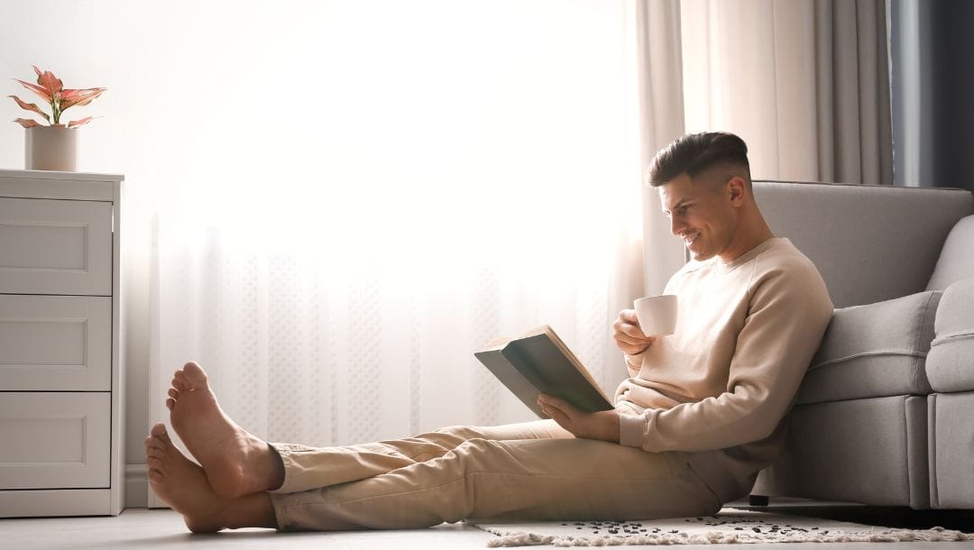 Smiling man seated on the floor, holding a coffee cup, propped against a couch, immersed in a book.