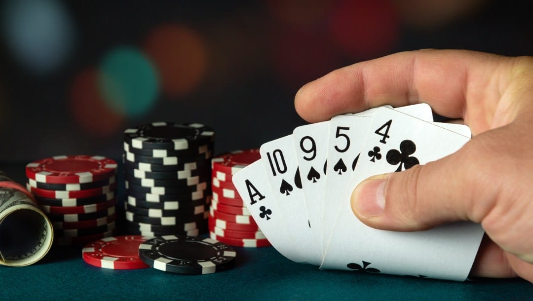 A hand holding five playing cards on a turquoise felt table. There are black and red casino chips with a roll of cash on the table.
