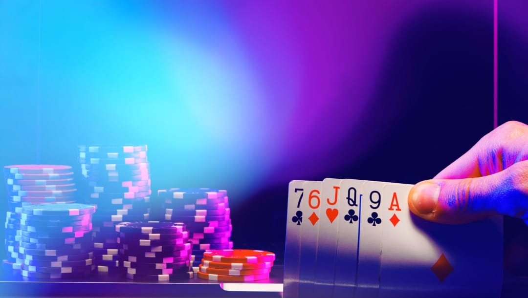 Fingers holding five playing cards. There is a laptop with casino chips on it. A blue and purple light is shining over the laptop screen.