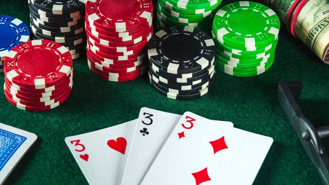 three of a kind three playing cards on a poker table surrounded by a deck of cards, poker chip stacks, rolled up money bills and a poker chip rake