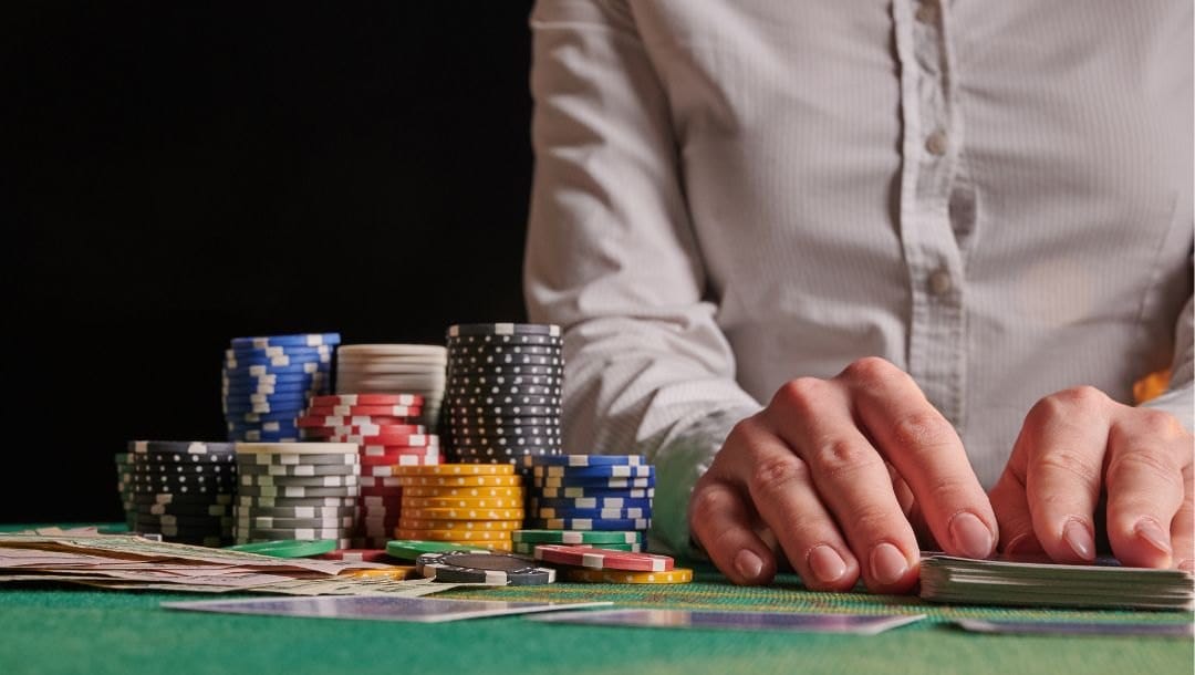 close up of a dealer’s hands resting on a pack of playing cards on a green felt poker table with poker chips stacked on it