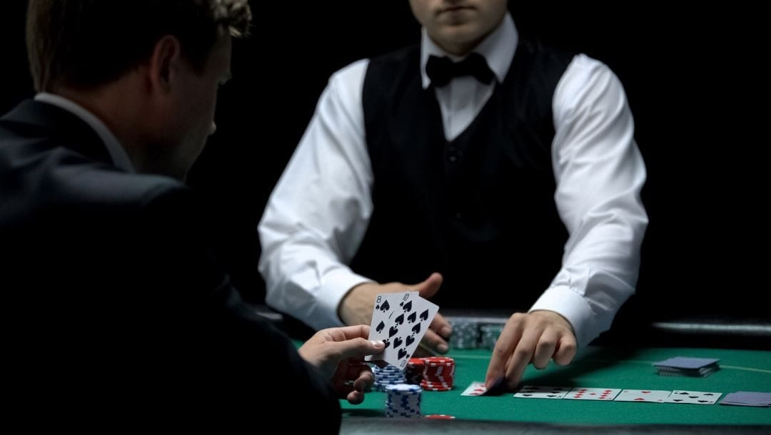 an over the shoulder view of a person holding two playing cards while sitting at a poker table as the dealer places another playing card on the table with poker chips on it