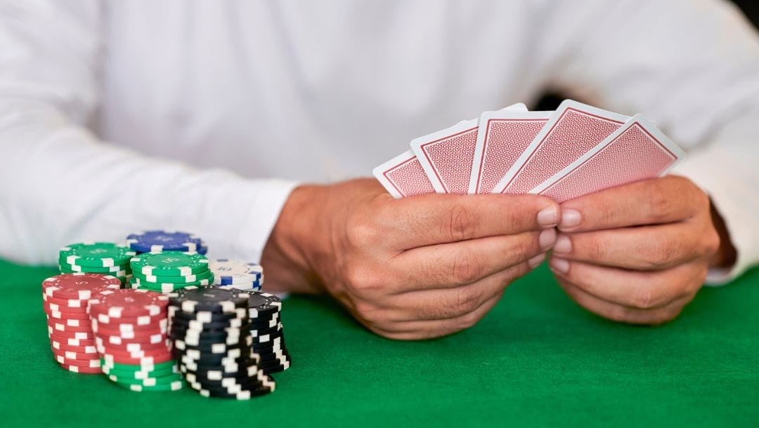 A person sitting at a poker table, holding playing cards, with stacks of red, white, blue, green, and black poker chips in front of him.