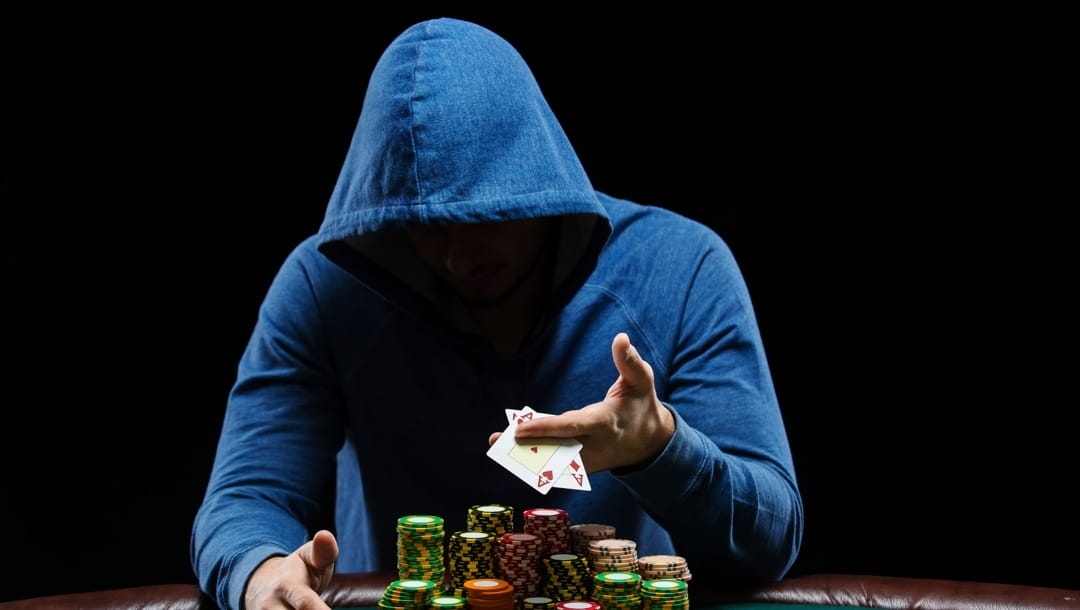 A faceless hooded man plays poker alone