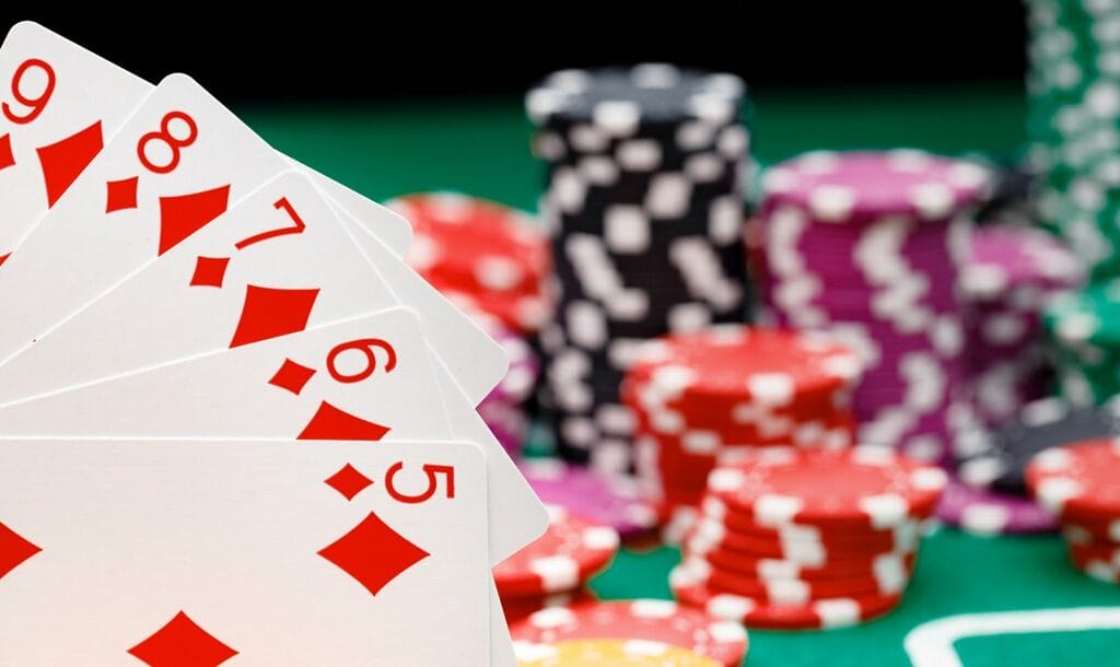 A straight flush held in front of stacks of poker chips on a poker table. The straight flush is the five to nine of diamonds.